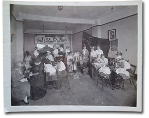 historical photo of several women in a room with a banner in the background that reads &ldquo;I.O.D.E Sock Shower for Soldiers Headquarters, 3rd floor, free wool to knitters.&rdquo; Many of the women are kitting or operating sewing machines, while others pour tea.