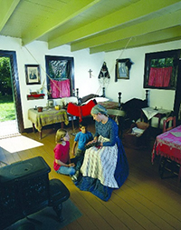 Interior of old house with a female interpreter dressed in period costume showing a M&eacute;tis sash to two children